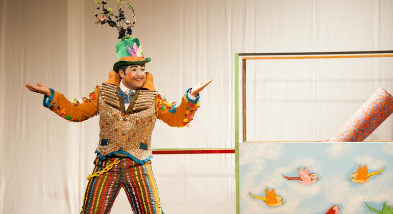 Ator com roupas estilizadas, casaco laranja e dourado calça colorida, e chapéu verde com uma planta em cima, com forte maquiagem, com estilo de palhaço, se apresenta no palco. 