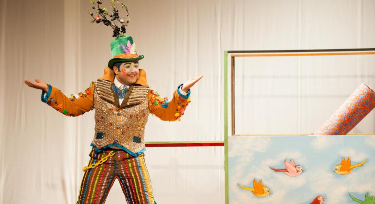 Peça aborda sustentabilidade no Teatro Raul Belém Machado. Foto: Alyson Jardin Ator com roupas estilizadas, casaco laranja e dourado calça colorida, e chapéu verde com uma planta em cima, com forte maquiagem, com estilo de palhaço, se apresenta no palco.