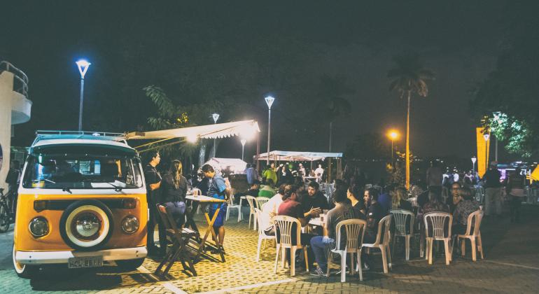 Pintura ao vivo e atrações musicais fazem parte da programação do Pampulha Noturno. Foto: Ricardo Laf Kombi amarela é espaço de oferta de alimentos na praça Dino Barbieri; mais de dez pessoas frequantam o local, sentadas em cadeiras e conversando ou em pé. Ao fundo, outras barracas de alimentos.