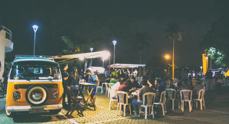 Kombi amarela é espaço de oferta de alimentos na praça Dino Barbieri; mais de dez pessoas frequantam o local, sentadas em cadeiras e conversando ou em pé. Ao fundo, outras barracas de alimentos. 