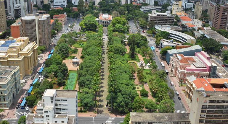 Vista aérea da Praça da Liberdade, com destaque para árvores, palmeiras e passeios e a rua central na qual se pode ver, ao fundo, o Palácio da Liberdade. Ao redor, prédios de mais diferentes estilos no entorno da praça.