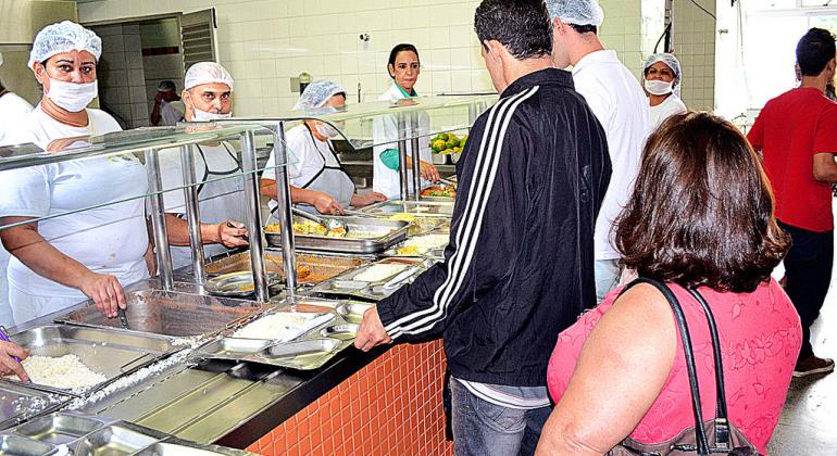 Conheça os cardápios dos restaurantes populares conforme os dias da semana. Foto: Denis Dias Dois cidadãos passam a bandeja e são servidos por funcionários do Restaurante Popular do Barreiro durante o dia.