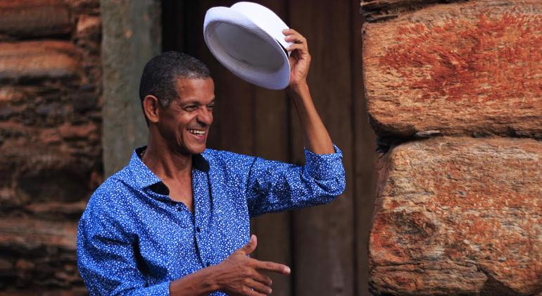 Foto de um senhor de blusa azul levantando um chapéu