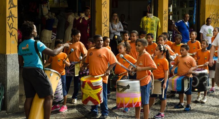 Crianças com camisetas cor laranja com instrumentos de percussão em frente à entrada Shopping Caetés