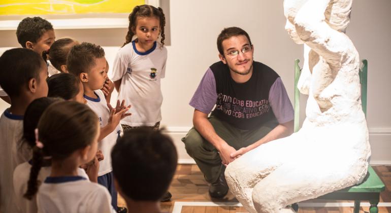 Projeto Circuito de Museus - Foto: Pablo Bernardo Na foto vê-se 9 crianças uniformizadas, de aproximadamente 7 anos, em uma sala de um museu, um professor agachado vestindo uma calça verde e uma camiseta preta com mangas roxas. Ele estão em frente á uma escultura toda branca de uma mulher nua, sentada em uma cadeira verde com o braço esquerdo levantado segurando a cabeça