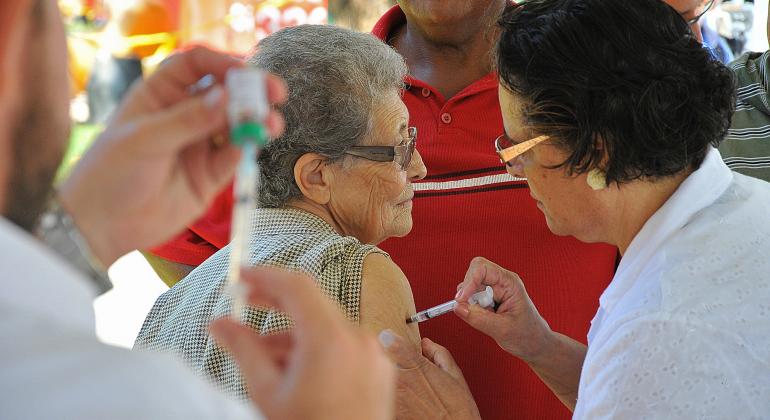 Campanha tem o objetivo de atingir 90% do público até dia 26 de maio. Foto: Breno Pataro Agente de saúde vacina idosa. Mais à frente, outro agente de saúde prepara aspira líquido da vacina com seringa.