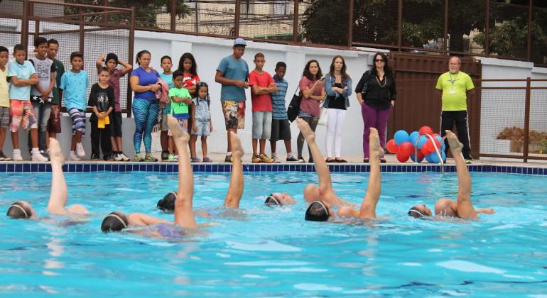 Piscina, com água bem azul, onde se passa uma apresentação de nado sincronizado de seis meninas com a perna direita fora d'água e as pontas dos pés esticadas. Do lado de fora, cerca de vinte pessoas olham para a água, dé pé. Há um amarrado de balões coloridos no canto do lado de fora da piscina.