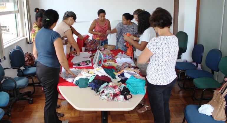 Cerca de sete mulheres em torno de uma mesa com várias roupas realizam oficina de convivência; 