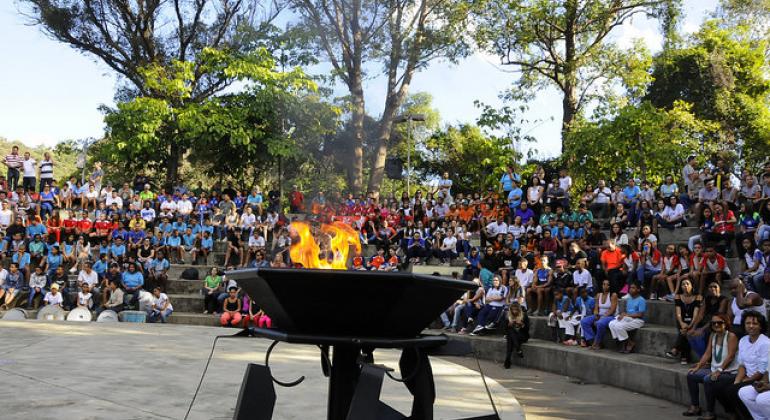 A foto mostra pessoas sentadas em arquibancadas de cimento, em um parque com árvores ao fundo. O céu está azul. A maior parte do público é constituída de alunos uniformizados. Em um plano superior, ao meio, está situada uma grande o pira em metal preto, e o fogo está aceso, uma chama grande e amarela.