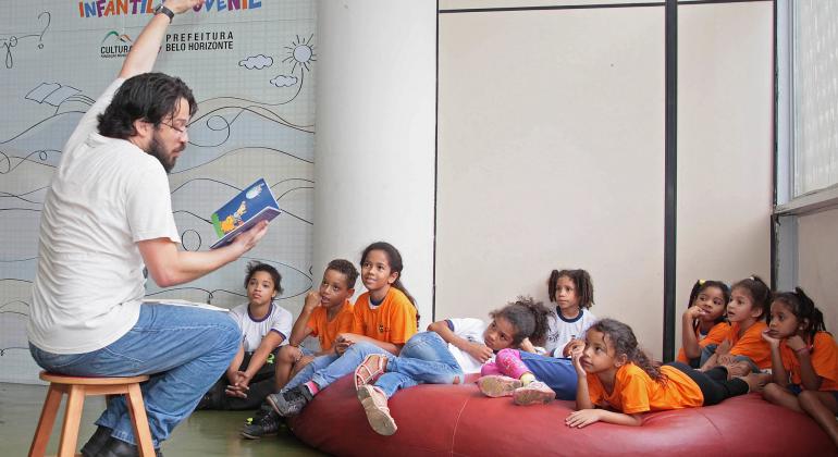 Contador de história lê para nove crianças, que estão sentadas em um grande colchão vermelho.