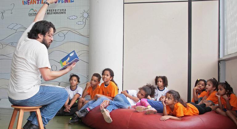 Encontro na Biblioteca Infantil e Juvenil debate o racismo - Foto: Divulgação/PBH Contador de história lê para nove crianças, que estão sentadas em um grande colchão vermelho.