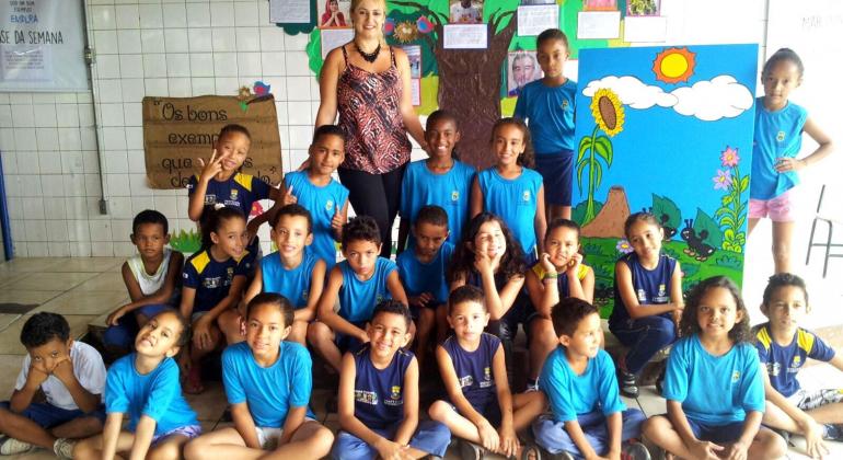 Crianças, estudantes da rede municipal de ensino, posam para a foto junto com professora. Ao fundo, painel de árvore.