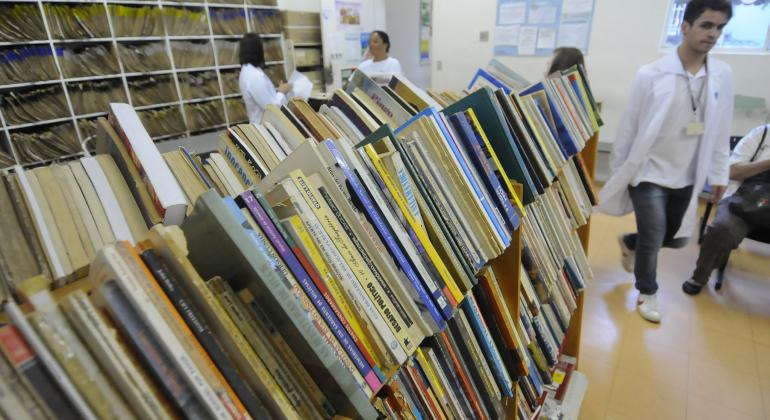 BH em Pauta: Uma graça de leitura - Foto: Rodrigo Clemente/PBH Estantes cheias de livros no primeiro plano. Ao fundo, duas mulheres de branco conversam. À direita, homem de jaleco faz movimento de caminhada.