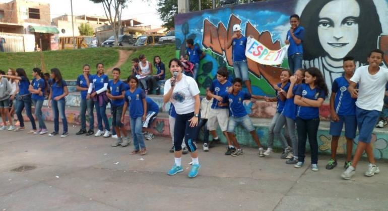 Crianças uniformizadas com blusas azuis da escola, e uma mulher falando no microfone posicionada na frente dessas crianças. 