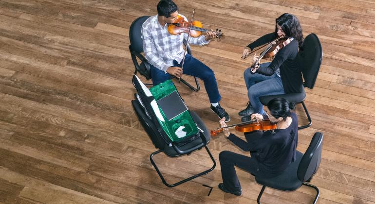 Três violinistas tocam, sentados em cadeiras. São quatro cadeiras e em uma delas 