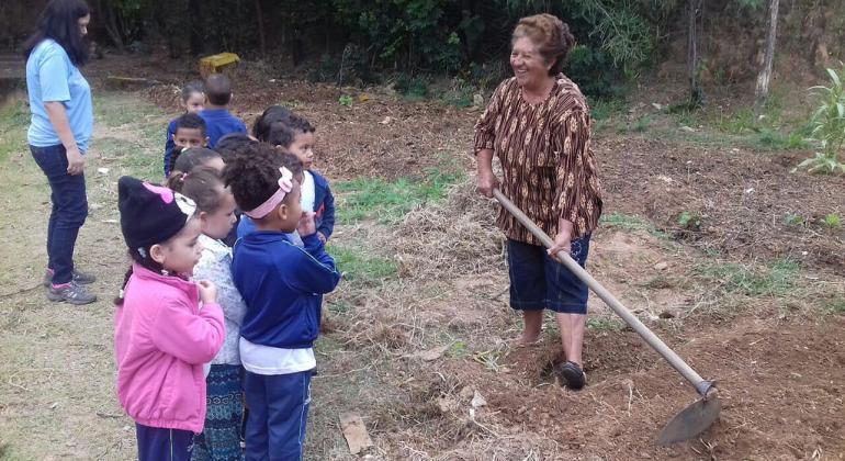 Crianças, estudantes da rede municipal de ensino, observam dona Maria da Gloria Silva, do grupo Amigas da Horta, que está com uma enxada na mão.