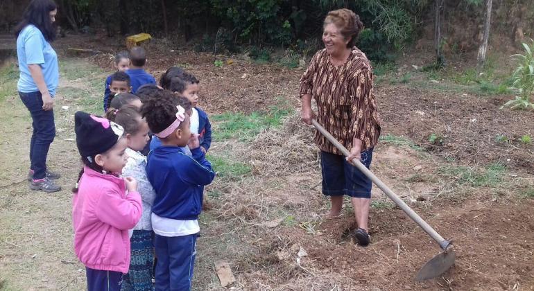BH em Pauta: Amigas da Horta Crianças, estudantes da rede municipal de ensino, observam dona Maria da Gloria Silva, do grupo Amigas da Horta, que está com uma enxada na mão.