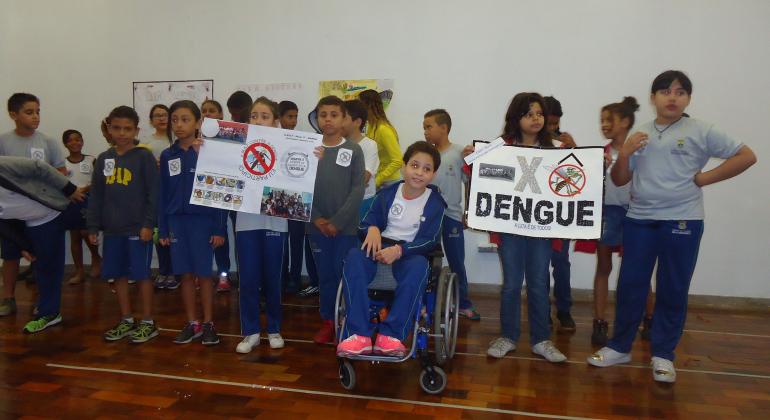 Cerca de 20 crianças, estudantes da rede municipal de ensino na foto. Uma delas está de em uma cadeira de rodas e as demais estão de pé. Uma delas segura um cartaz com um símbolo de negativo (X) e um escrito "dengue".