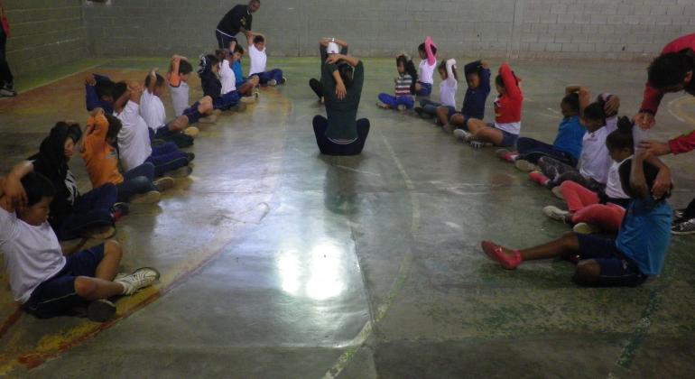 Crianças sentadas em meio circulo e fazendo alongamentos.