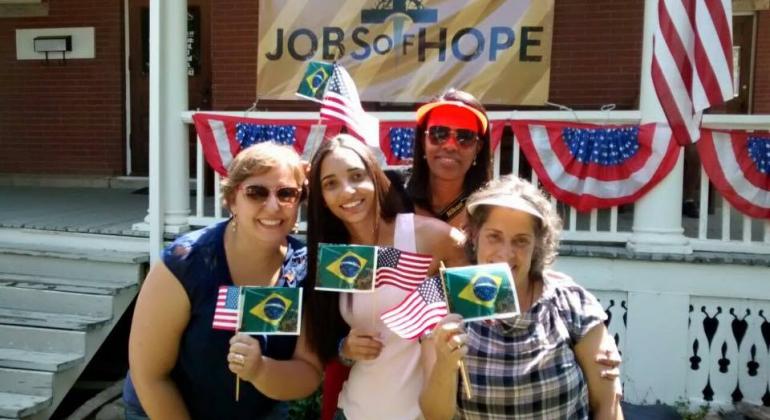 Professoras da rede municipal de ensino participam de celebração do 4 de julho nos Estados Unidos. Na foto, 4 mulheres seguram bandeirinhas do Brasil e dos Estados Unidos