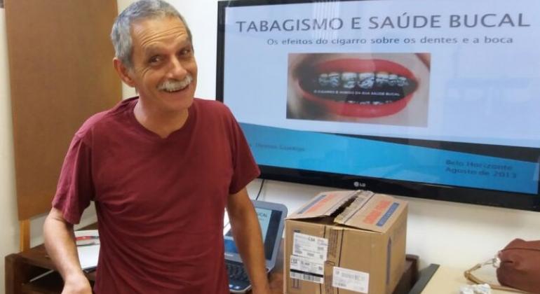 Homem sorri na foto. Ao fundo, tela com texto "Tabagismo e saúde bucal"