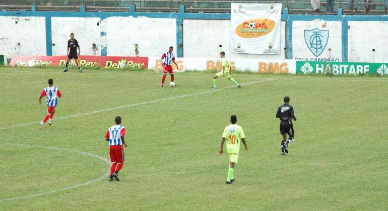 Na foto, cinco jogadores do futebol masculino e o árbitro da partida. Campo de grama. Ao fundo, bandeira com o símbolo da Copa Centenário.