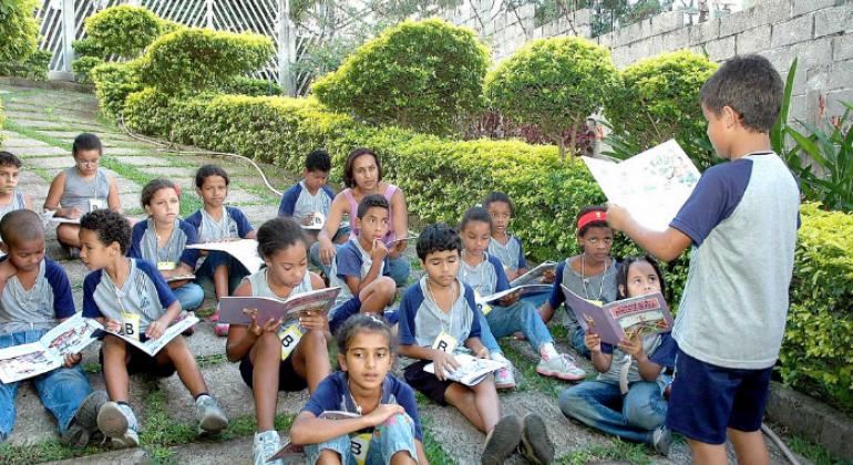 Crianças, estudantes da rede municipal de ensino, observam, sentados, o colega que lê de pé. Ao fundo, o jardim da escola.