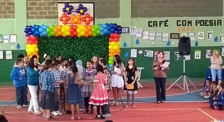 Mais de dez alunos con trajes de festa junina se organizem em frente a palco para se apresentarem. Ao lado, professora e ao fundo, os Dizeres "Xafé com poesia". 