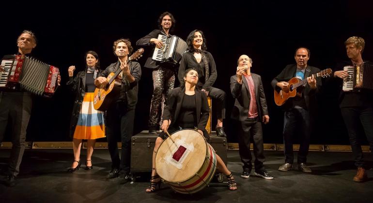 Grupo de oito atores do Grupo Galpão posam com instrumentos musicais. Dois seguras sanfonas, outros dois, violões. Uma está com tambor. Todos vestem preto.