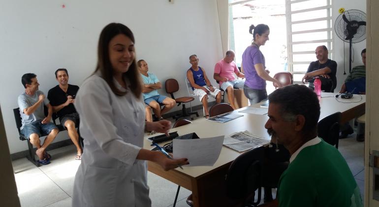 Centro de Saúde promove o Dia de Atenção à Saúde do Homem - Foto: Divulgação/PBH No primeiro plano, funcionária da saúde segura folha de exames de paciente. Ao fundo, fila com outros pacientes que aguardam atendimento. Todos são homens.
