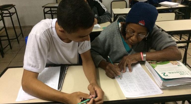 Jovem aluno e mulher de idade que fazem parte do Programa de Educação de Jovens e Adultos do Barreiro estudam sentados.