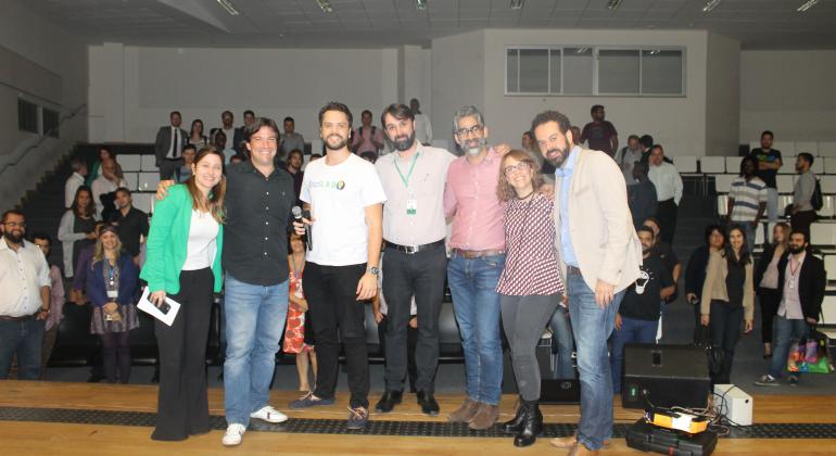 Painelistas e representantes da Prefeitura e BrazilLAB posam para foto, registrada no palco, tendo ao fundo os participantes do evento