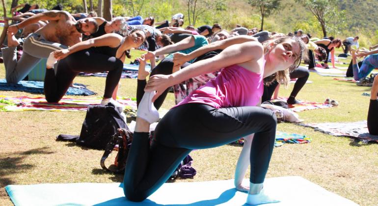 Ioga pode ser praticada gratuitamente em alguns parques municipais de Belo Horizonte. Foto: FPM/PBH Mais de dez pessoas fazem pose de Ioga em gramado de parque durante o dia.