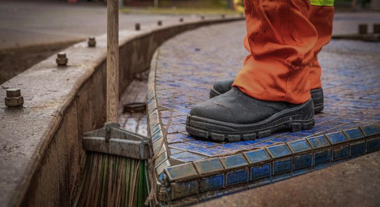 Foto com recorte no pé de um gari, que usa bota, enquanto limpa, com vassoura, fonte na savassi