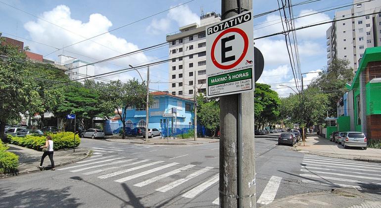 Cruzamento de trânsito com placa de Estacionamento Rotativo em uma das esquinas, durante o dia.