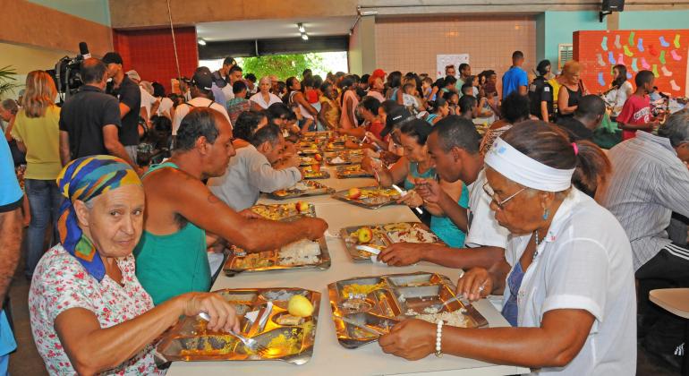 Mais de trenta pessoas das mais diversas idades almoçam em um Restaurante Popular. 