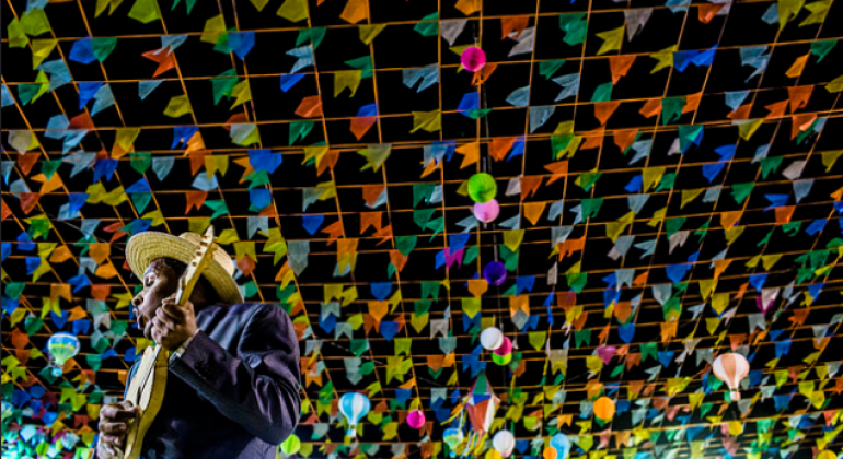 Prefeitura divulga shows do Arraial de Belo Horizonte - Foto: Samuel Mendes Quadrilheiro toca viola. Ao fundo, muitas bandeirinhas. Foto é um contra-plongé.