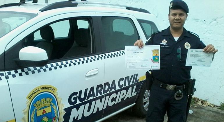 Capacitação para os guardas municipais em cursos de idiomas Guarda Municipal, ao lado de uma viatura, segura diplomas de cursos de capacitação em línguas
