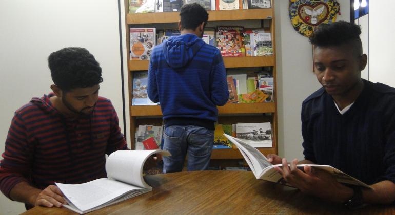 dois adolescentes sentados em uma mesa lendo um livro, um outro adolescente em pé de frente para uma estante de livros. 