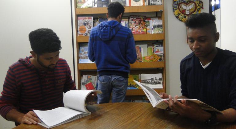 Superintendência de Limpeza Urbana é fonte de pesquisa para estudantes e empresas; mais de mil atendimentos são realizados por ano. Foto: Pedro Antônio de Oliveira/PBH dois adolescentes sentados em uma mesa lendo um livro, um outro adolescente em pé de frente para uma estante de livros.