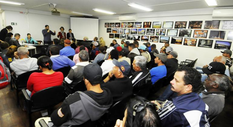 Resultado do sorteio das vagas para camelôs - Foto: Adão Souza/PBH Cerca de 40 pessoas sentadas em cadeiras pretas em um auditório. À frente, no pequeno palco, três pessoas fazem um sorteio