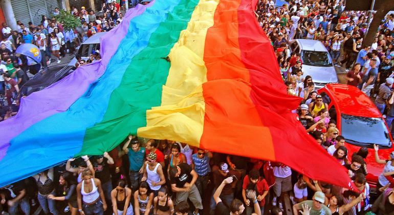 Mais de cem pessoas seguram a bandeira do Arco-íris, símbolo da luta LGBT, em avenida, com carros estacionados. 