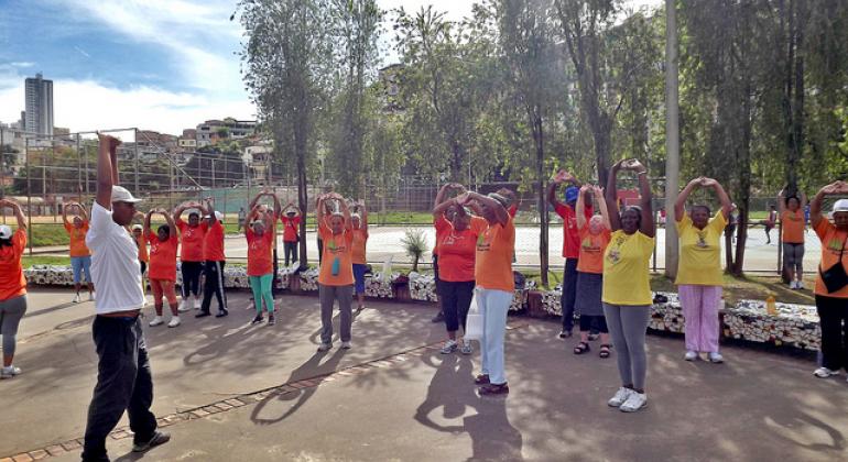 Os selecionados atuarão nos programas desenvolvidos pela Secretaria Municipal de Esporte e Lazer. Foto: SMEL/PBH Numa área aberta, professor faz exercício de alongamento junto a alunos da terceira idade.