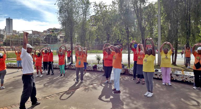 Numa área aberta, professor faz exercício de alongamento junto a alunos da terceira idade.