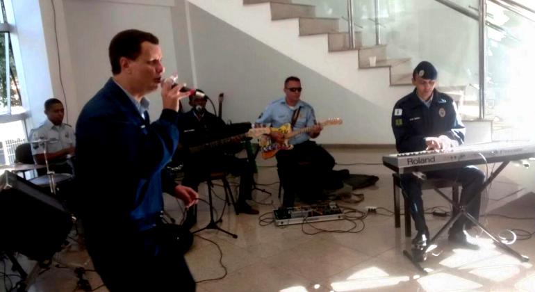 Com repertório eclético e dançante, grupo musical da Guarda Municipal anima eventos públicos, privados e religiosos. Foto: COP/PBH Cinco membros da banda da Guarda Municipal se apresentam em ambiente interno durante o dia. Além do vocalista, estão presentes membros utilizando instrumentos como órgão e guitarra.