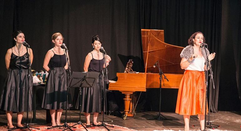 No palco, atrizes encenam o musical "As Rainhas do Rádio": uma está cantando à frente de um piano de cauda e, ao lado dela, três mulheres fazem "baking vocal".