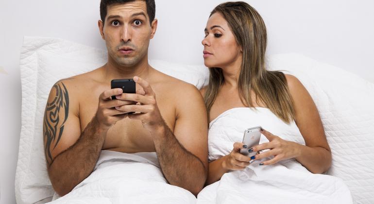 Foto promocional da peça com os dois atores utilizando o celular deitados em uma cama; mulher espia o celular do homem. Foto: Nello Aun