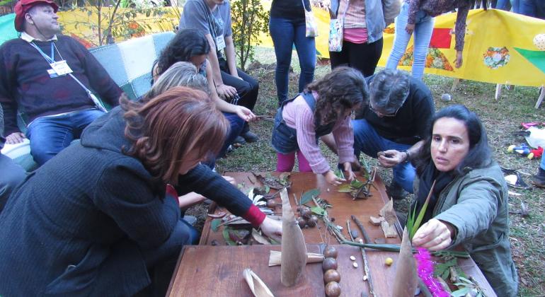 Cerca de dez pessoas de idades diversas participam de oficina de brinquedos no chão manipulando gravetos e folhas em uma mesa, em uma área verde, durante o dia.