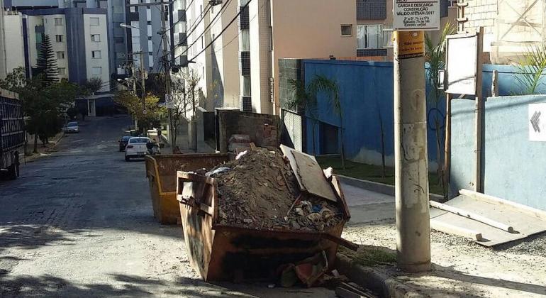 Duas caçambas cheias de entulho próximas à calçada de uma rua de BH durante o dia. 