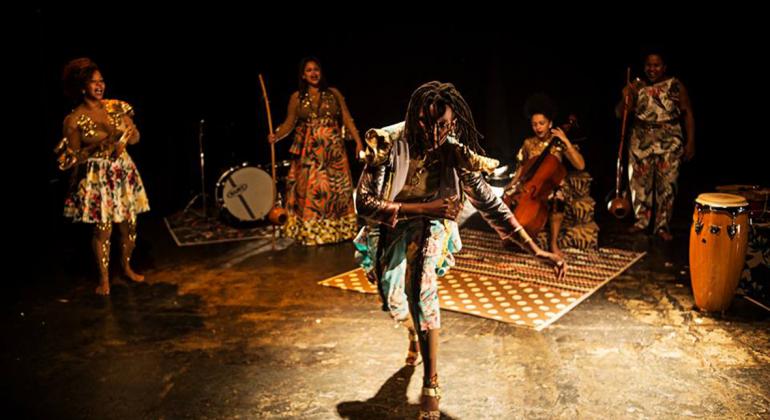 Mulher negra com tranças no cabelo dança. Ao fundo, três mulheres negras com instrumentos musicais tocam em cenário com inspiração africana. Peça Eras, do Coletivo Negras Autoras